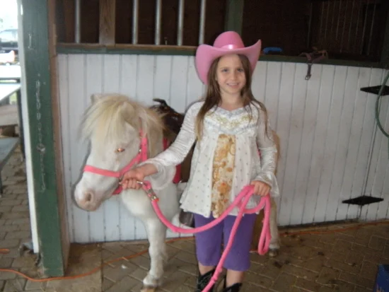 A smiling child in a pink cowboy hat beside a pony